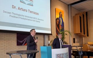 Links een man in pak met microfoon in de hand, rechts een man in pak achter een katheder.