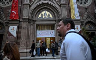 Mensen komen uit een grote kerk, met op de deur een banner, die ”Pope Leo XIV” welkom in Turkije heet.