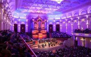 Blauw verlicht Concertgebouw met op het podium een koor en orkest met dirigent. Daarachter een enorm orgel en rijen vol luisteraars om het podium heen.