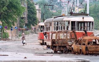 Een oude vrouw loopt over straat. Naast haar staan uitgebrande auto’s en een kapotte tram. 