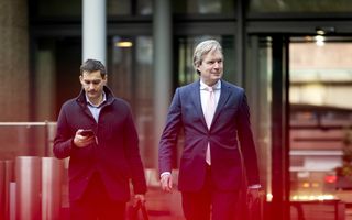 Man in pak loopt buiten voor een gebouw. Aan zijn rechterzijde loopt een man met een telefoon in zijn hand.
