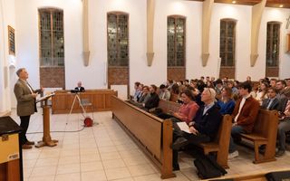 Een man achter een katheder, links, spreekt -met name- jonge mensen in de kerk toe.