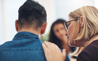 Blonde vrouw met bril op legt haar hand op de schouder van een man met een blauw overhemd. Op de achtergrond kijkt een meisje toe.