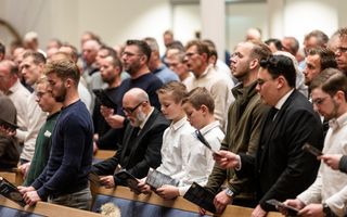 Mannen en jongens in nette kleding staan in kerkbanken te zingen.