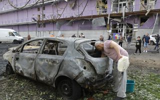 Een man in roze shirt en met een tas in zijn hand bekijkt een uitgebrande auto. Op de achtergrond een beschadigd complex.