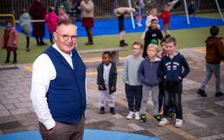 Een oudere man met ronde bril, grijze broek en blauw giletje staat op een plein met schoolkinderen.