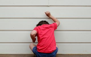 Jongen met rood shirt aan en blauwe korte broek zit met zijn gezicht naar een muur gekeerd. Met zijn hoofd en met zijn vuist bonkt hij tegen de muur.