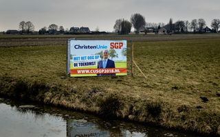 Een verkiezingsbord in een weiland, langs water. De namen van ChristenUnie en SGP staan er onder meer op. 