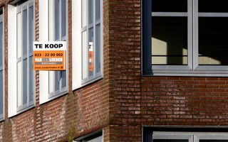 Deel van een appartementencomplex. Daarop een oranje met wit bord waarop staat aangegeven dat een woning te koop is.