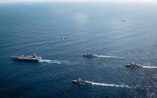 Luchtfoto van de oceaan met daarop een groot vliegdekschip en drie kleinere marineschepen. In de lucht vliegen bommenwerpers en jachtvliegtuigen.