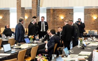 Een stuk of tien mannen in donkere pakken staan of zitten aan vergadertafels en spreken met elkaar.