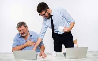 Oudere man kijkt naar zijn laptop. Een jongere man staat naast hem en wijst op de laptop van de oudere man iets aan. 