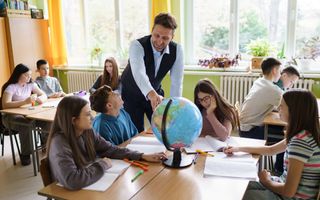 Leraar staat bij een groepje tafeltjes in klaslokaal en vertelt iets over de wereldbol ie op tafel staat. Leerlingen luisteren.
