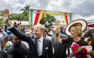 Man in pak en vrouw met hoed zwaaien terwijl ze tussen een menigte mensen lopen.