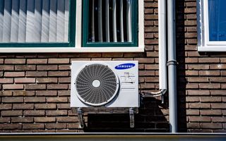 Een wit kastje met daarin een grote ventilator hangt aan een bakstenen muur met rechts ernaast een regenpijp.