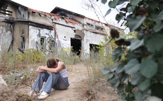 Vrouw zit met hoofd op haar armen op de grond voor zwartgeblakerde huizen.