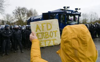 Een persoon gehuld in een gele cape houdt een bord omhoog met de tekst: AfD verbot jetzt. Op de achtergrond staan politieagenten. 