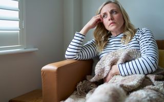 Een zieke vrouw zit op de bank met een kleed over haar heen. 