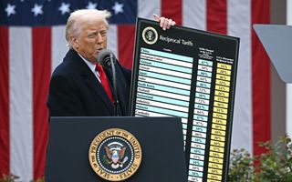Een oudere man met grijs haar en een rode stropdas houdt een bord omhoog met namen van landen en lijsten percentages. Op de achtergrond een grote Amerikaanse vlag.