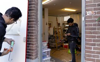 Man spuit kit rond een brievenbusopening in een deur; door de deuropening zie je een woonkamer waar iemand met een tochtstrip bezig is.