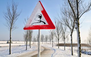 Driehoekig waarschuwingsbord voor een gladde weg staat langs een weg met een bomenlaan bedekt door sneeuw en ijs.