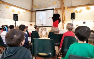 Kinderen op een zolder van een school, zittend op stoeltjes, luisteren naar een juf op een podium. Het podium is versierd met gele sterren. Er is een scherm en er staan geluidsboxen. 