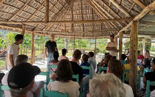 Mensen onder een rieten overkapping. Ze luisteren naar een man die staat.