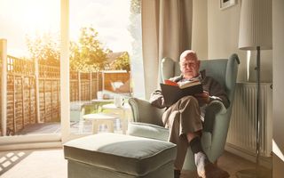 Een oudere man zit in een zonnige kamer in een stoel een boek te lezen. 