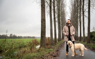 Man van middelbare leeftijd in beige jas en zwarte broek staat buiten op een asfaltpaadje met een blonde hond. Aan weerszijden van het pad staan bomen. 