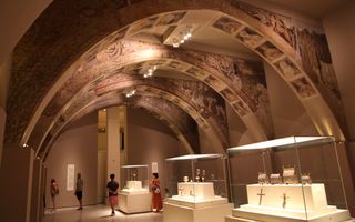 Museumzaal met aan het plafond bogen met daarop muurschilderingen. Mensen lopen langs enkele vitrines met voorwerpen.