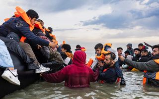 Donkere mensen met oranje hesjes staan in zee in een poging aan boord van een opblaasbootje te gaan. 