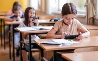 Meisje met beige shirt aan tafeltje in schoollokaal met smartphone in haar hand.