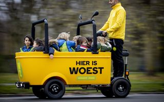 Een man met een geel hesje staat achterop een knalgele elektrische kar met daarin zo’n acht kinderen.