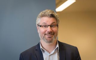 Portret van man met baard en donkere bril voor een blauwe en gele wand.