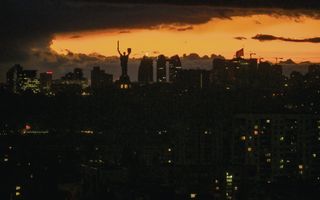 Een grotendeels donkere stad, met op de achtergrond een oranje lucht. Op een van de gebouwen staat een groot beeld met een geheven zwaard en schild. 