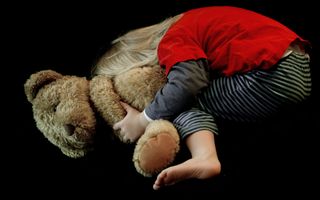 Een jong kind met een rood shirtje en gestreepte broek aan duikt weg achter een grote knuffelbeer.