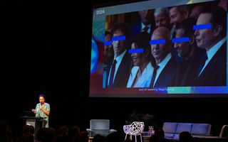 Vrouw in lichtgroene jurk houdt microfoon vast. Op het grote scherm achter haar zijn zes personen te zien met blauwe balkjes voor hun ogen. 