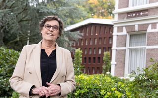 Vrouw met donker haar, bril, kruisje om haar hals, licht jasje.