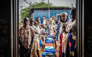 Vrouwen met kinderen staan in twee rijen te wachten voor de ingang van een huis. Tussen hen in staat een rij met gekleurde emmers.
