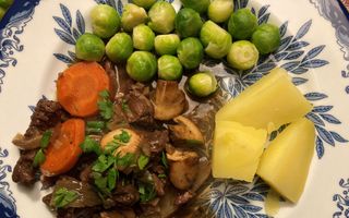 Op een crème bord met blauwe bloemen liggen stukjes rundvlees in een saus. Ernaast spruitjes, schijfjes wortel en stukjes aardappel.