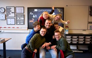 Een man in een blauwe trui zit op een stoel in een klaslokaal. Naast hem zitten en staan zes leerlingen, dicht tegen hem aan. De twee meisjes die achter hem staan steken hun handen in de lucht. 