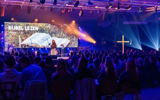 Donkere zaal vol jonge mensen, op de rug gezien. Op het podium staat een vrouw voor een scherm met daarop een opengeslagen Bijbel.  