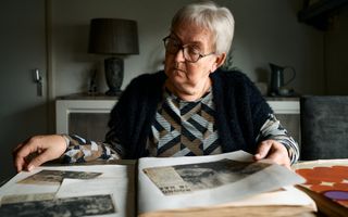 Een vrouw met wit haar en een bril bladert in een boek met krantenknipsels.