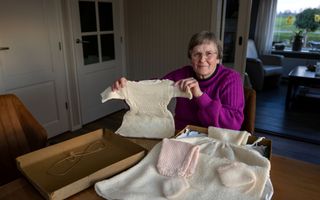 Mevrouw met paarse trui houdt babyjurkje omhoog. Op tafel voor haar een doos met nog meer babykleertjes.