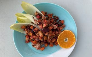 Lichtblauw bord waarop in kleine stukjes gesneden groenten liggen. Ernaast een schijfje sinaasappel en vier witlofblaadjes.