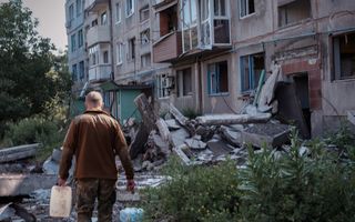 Een man met een bruine jas loopt met een jerrycan door een kapotgeschoten straat met kapotgeschoten flat