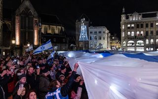 Een menigte houdt een reusachtige wit met blauwe vlag omhoog. Het is donker. Op de achtergrond verlichte gebouwen.