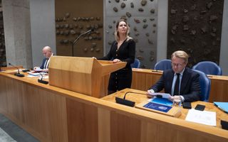 Vrouw staat achter spreekgestoelte in de Tweede Kamer. Aan beide zijden van haar zit een man.
