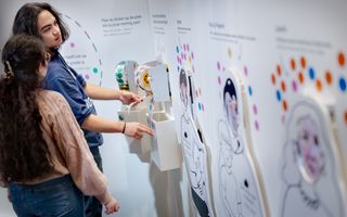 Een jongeman en een jonge vrouw staan bij een rol met stickers. Op de witte muur zijn al diverse stickers geplakt. De tekst die erbij staat, is niet goed zichtbaar.