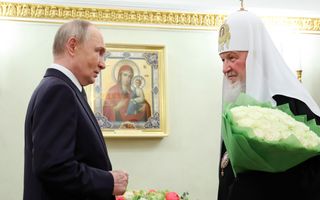 Een man in pak en een man in zwart ambtsgewaad met witte mijter met doek, met een bos witte rozen in de arm, kijken elkaar recht in het gezicht. Tussen hen in is een schilderij van een heilige te zien.